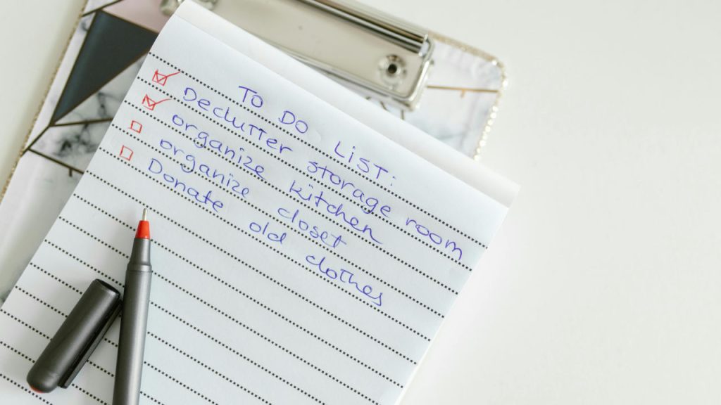 Notebook with a handwritten checklist, black pen placed on top, eyeglasses resting nearby, white background, organized task list format, red bullet points indicating completed or pending tasks