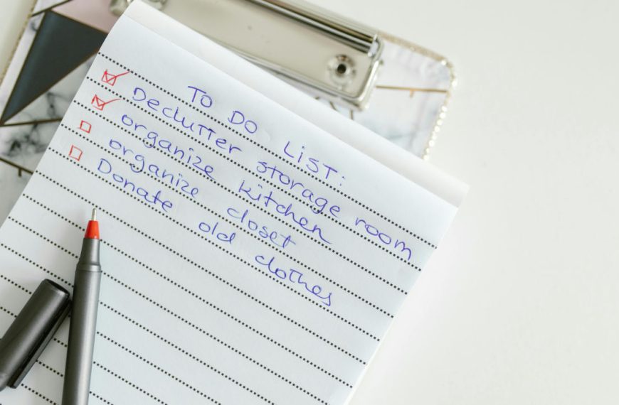 Notebook with a handwritten checklist, black pen placed on top, eyeglasses resting nearby, white background, organized task list format, red bullet points indicating completed or pending tasks