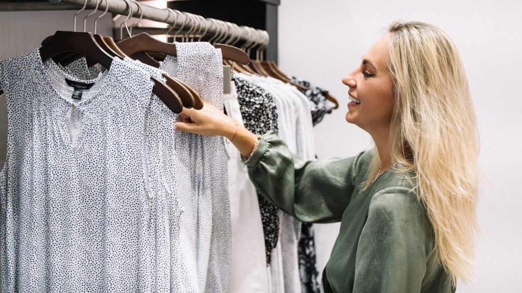 Woman choosing clothes from a rack, blonde hair, green blouse, selecting between white and light-colored shirts, standing in a modern wardrobe or closet