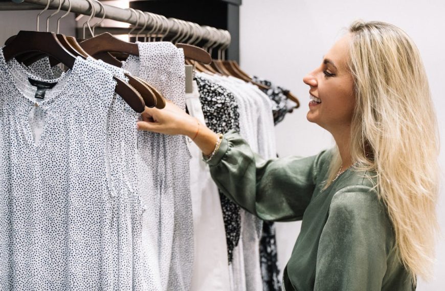 Woman choosing clothes from a rack, blonde hair, green blouse, selecting between white and light-colored shirts, standing in a modern wardrobe or closet