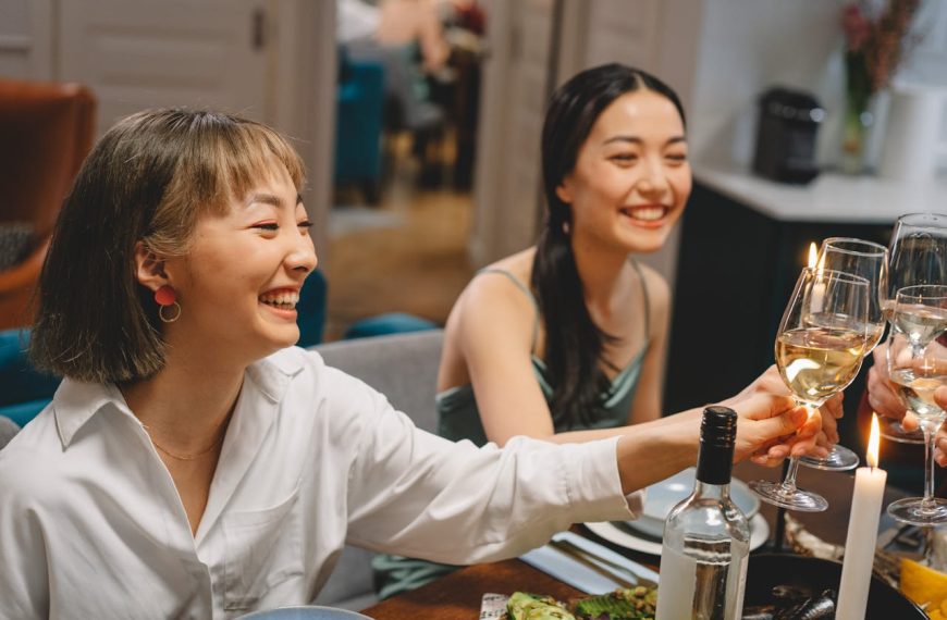 Two smiling women sitting at a dining table, raising glasses in a toast, with a bottle of wine and food visible in front of them