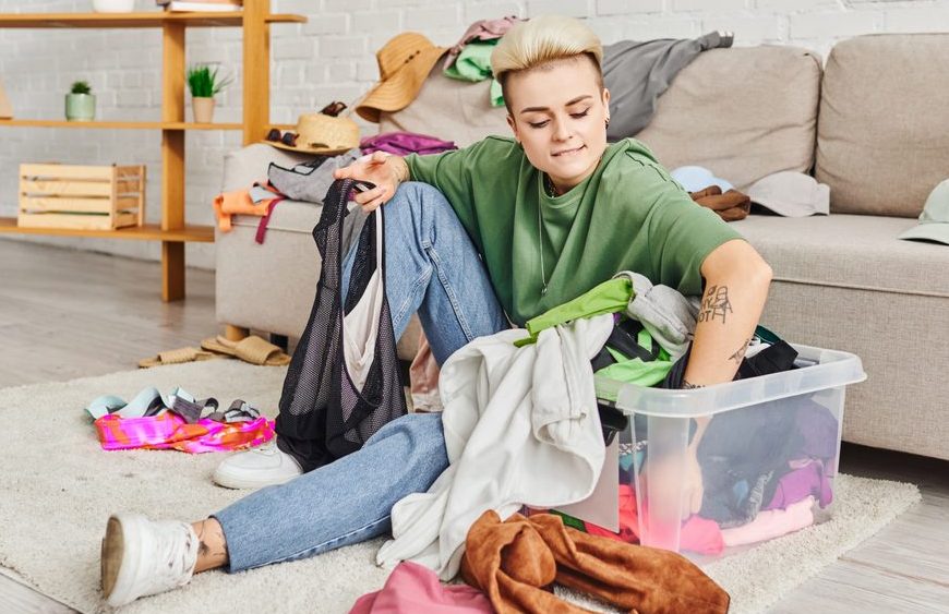 Person sitting on the floor sorting through a plastic bin of clothes, messy room with scattered clothing, couch and shelves in the background