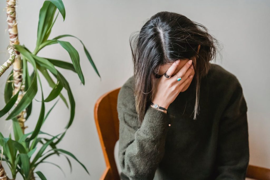 woman in green sweater sitting on chair, head in hand, fingers covering face, distressed posture, indoor plant in background