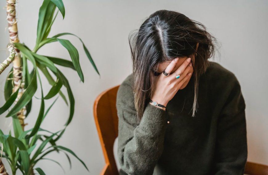 woman in green sweater sitting on chair, head in hand, fingers covering face, distressed posture, indoor plant in background