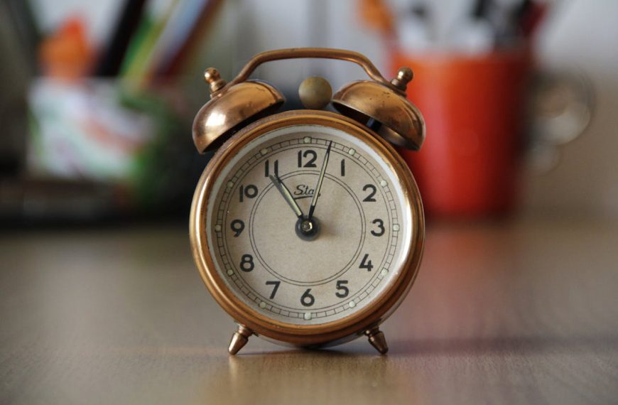 Vintage copper alarm clock on wooden surface, showing 7:00, blurred colorful background
