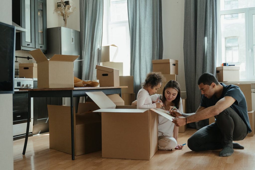 Family sitting on floor, unpacking boxes, child playing, surrounded by moving boxes, bright living room