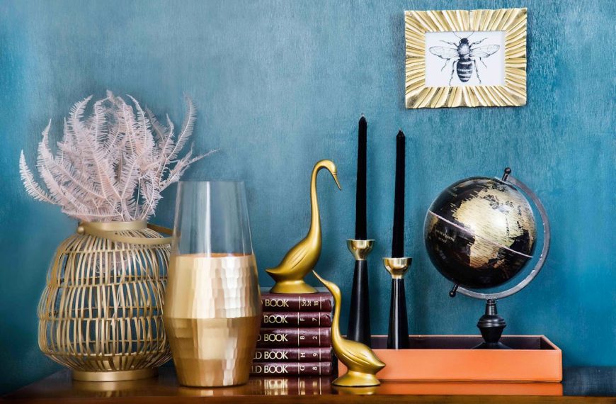 Elegant home decor arrangement, blue textured wall, gold and black decorative items, globe, swan figurines, stacked books, tall black candles, gold picture frame