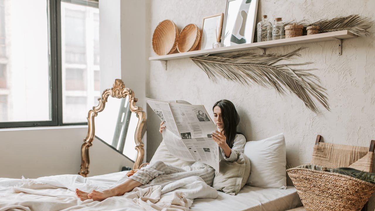 Woman sitting on bed reading newspaper, cozy bedroom setting, large mirror on the floor, decorative shelf with baskets and art, soft natural lighting