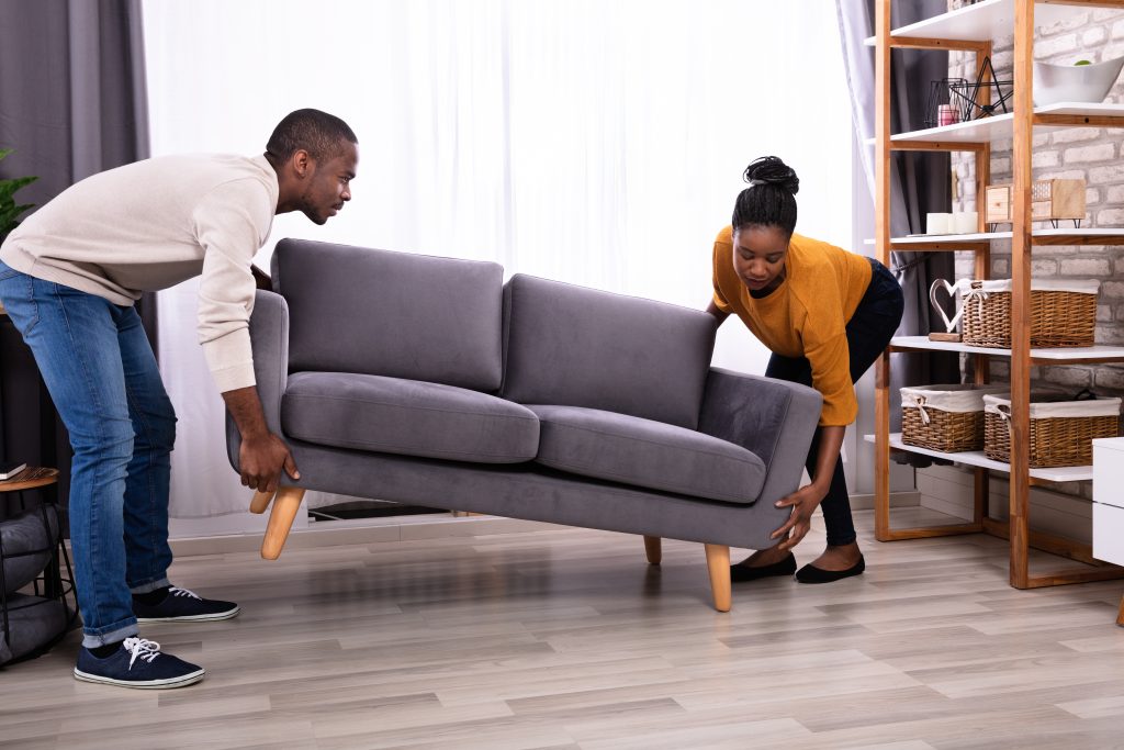 couple arranging furniture, man and woman lifting a couch, modern living room, light-colored walls, moving boxes nearby, teamwork, casual clothing, natural daylight
