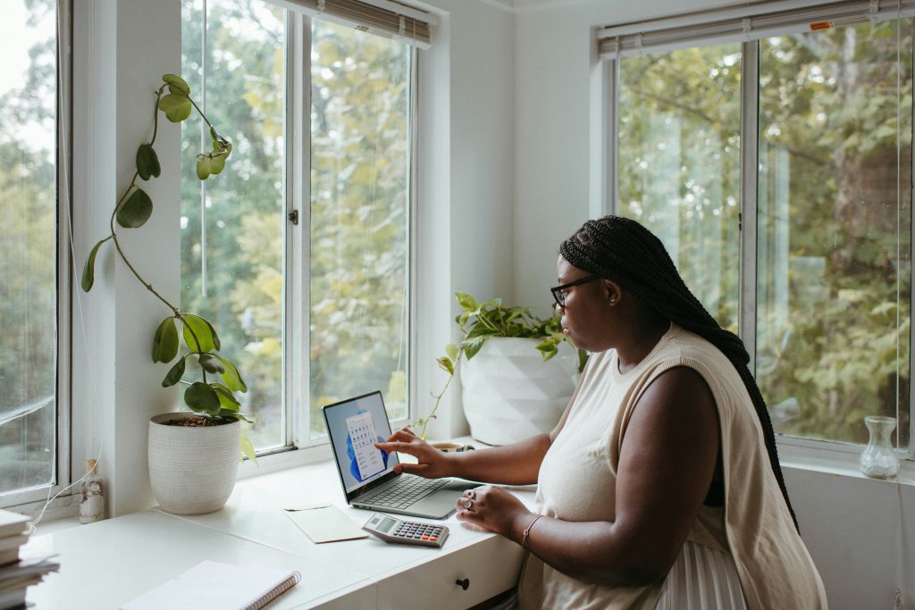 Woman working on a laptop, seated at a white desk, surrounded by green indoor plants, large windows letting in natural light, peaceful home office setting, daytime, focused expression, cozy and bright workspace