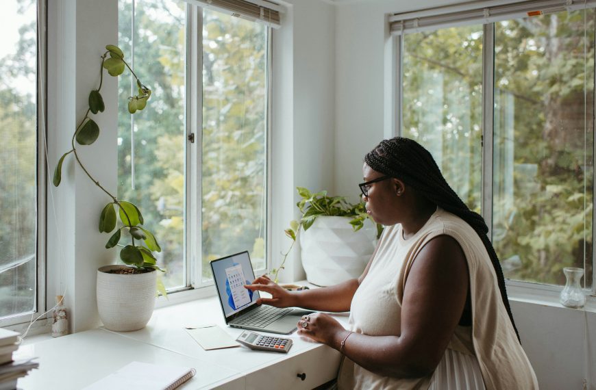 Woman working on a laptop, seated at a white desk, surrounded by green indoor plants, large windows letting in natural light, peaceful home office setting, daytime, focused expression, cozy and bright workspace