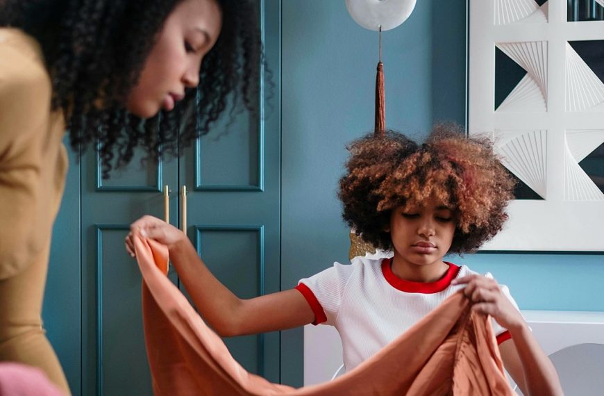 A young child with curly hair in a white shirt stands at a door, holding what appears to be paper or an envelope. An adult figure is partially visible in the background near a blue door