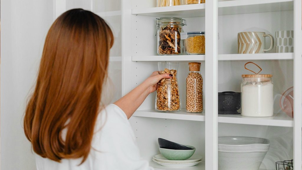 Woman organizing kitchen pantry, reaching for a jar of nuts, neatly arranged shelves with containers and dishes, modern kitchen storage, home organization, minimalistic interior design