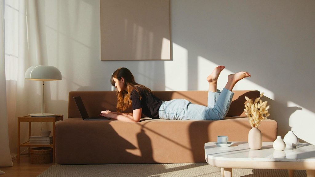 Woman lying on a brown sofa, using a laptop, sunlight streaming through the window, cozy living room, relaxed atmosphere, decorative table