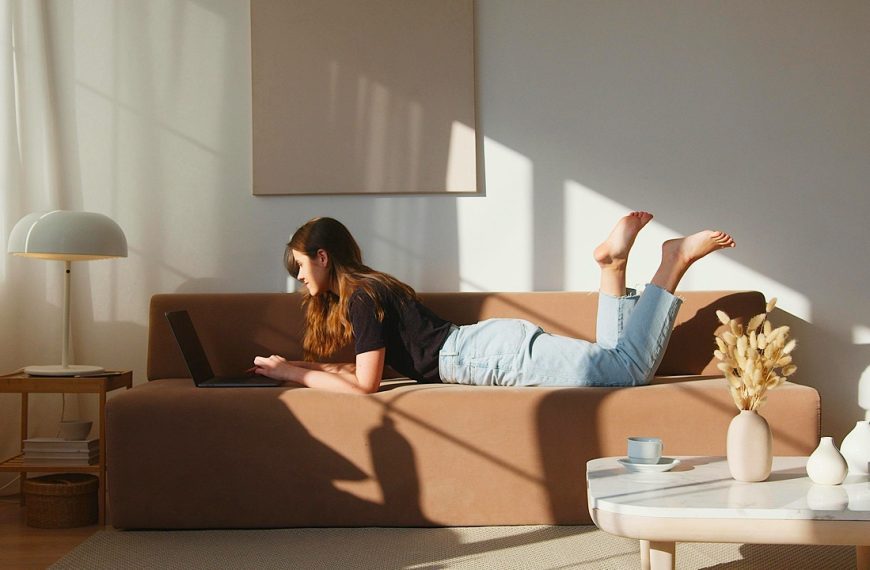 Woman lying on a brown sofa, using a laptop, sunlight streaming through the window, cozy living room, relaxed atmosphere, decorative table