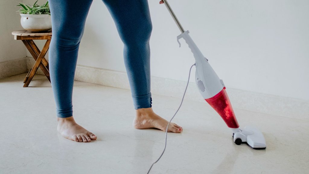 Person vacuuming a white floor, using a lightweight corded vacuum, barefoot, wearing blue jeans, home cleaning
