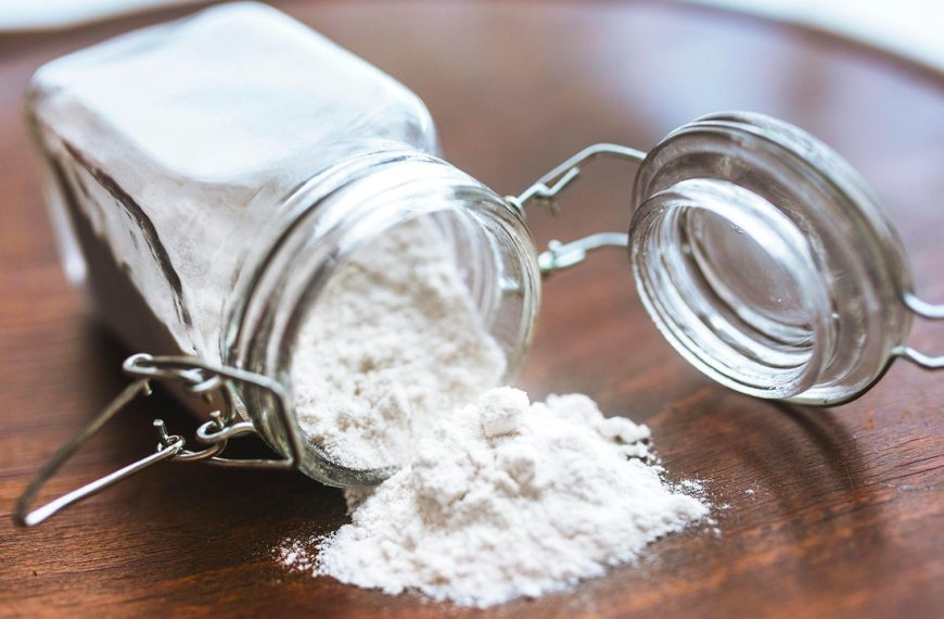 Glass jar tipped over with white flour spilling onto a wooden surface, metal clasp and lid open, fine powder scattered, baking ingredient in a kitchen setting