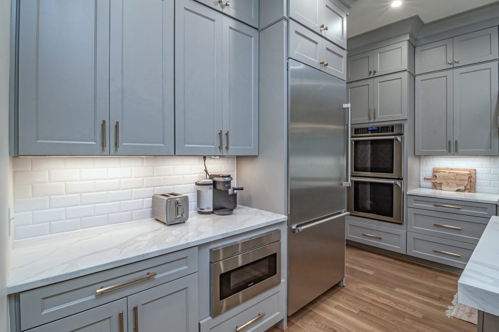 `Modern kitchen with light gray cabinetry, stainless steel refrigerator, built-in microwave, double wall oven, white subway tile backsplash, marble countertops, wooden flooring, under-cabinet lighting