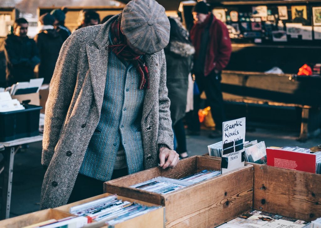 Man browsing vinyl records at an outdoor market, dressed in a gray coat, flat cap, and scarf, standing in front of a wooden box