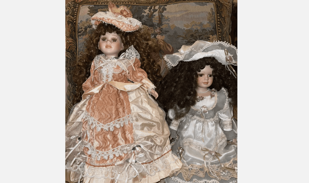 Two porcelain dolls with curly brown hair, dressed in detailed Victorian-style dresses, sitting on patterned cushion with tapestry background