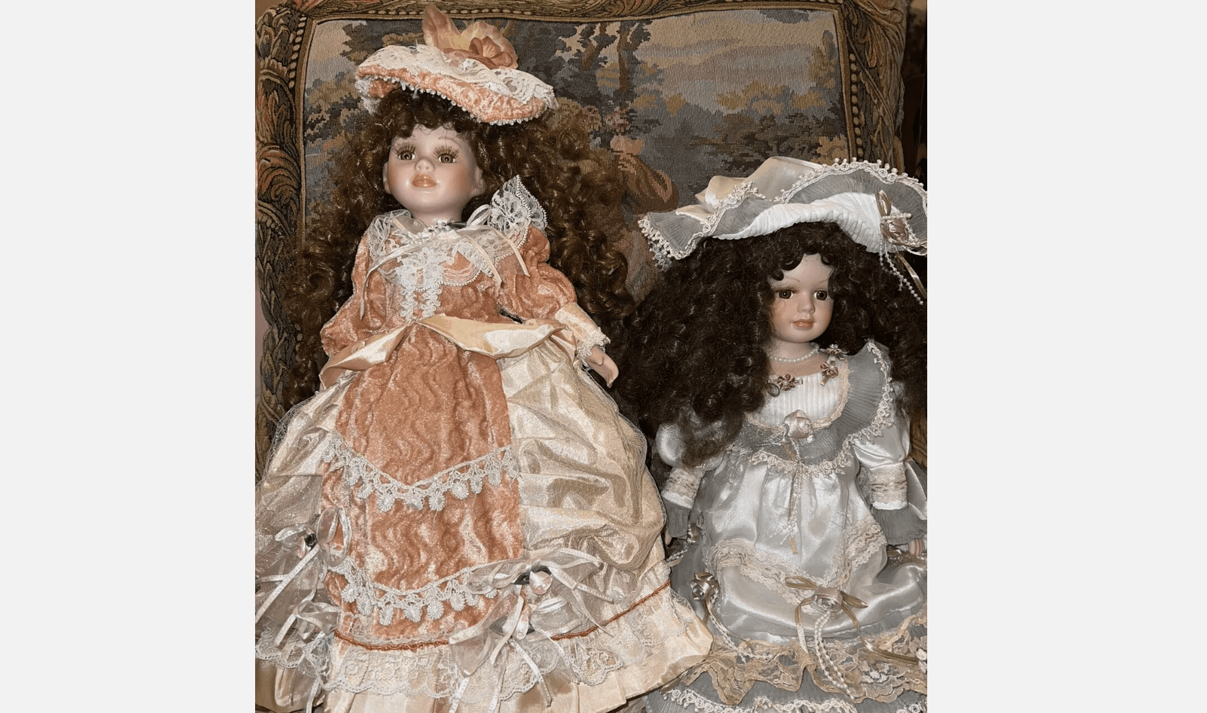 Two porcelain dolls with curly brown hair, dressed in detailed Victorian-style dresses, sitting on patterned cushion with tapestry background