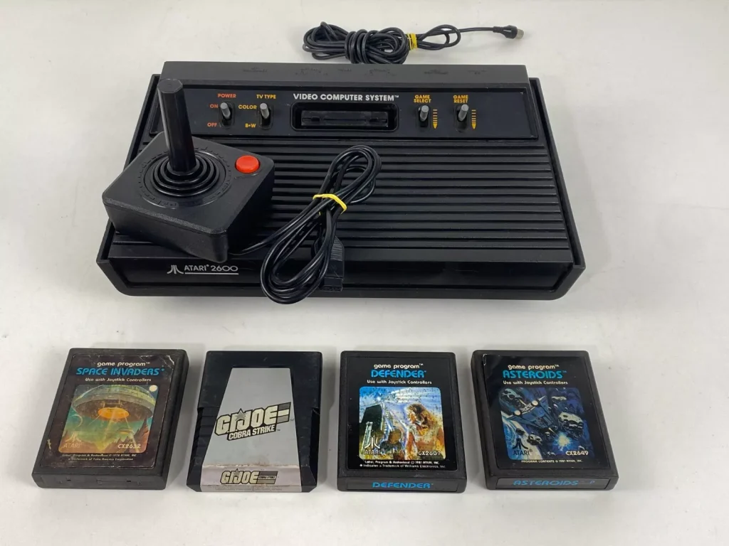Atari 2600 console, black design with ridged surface, classic joystick controller with a red button, four game cartridges displayed in front, vintage gaming system