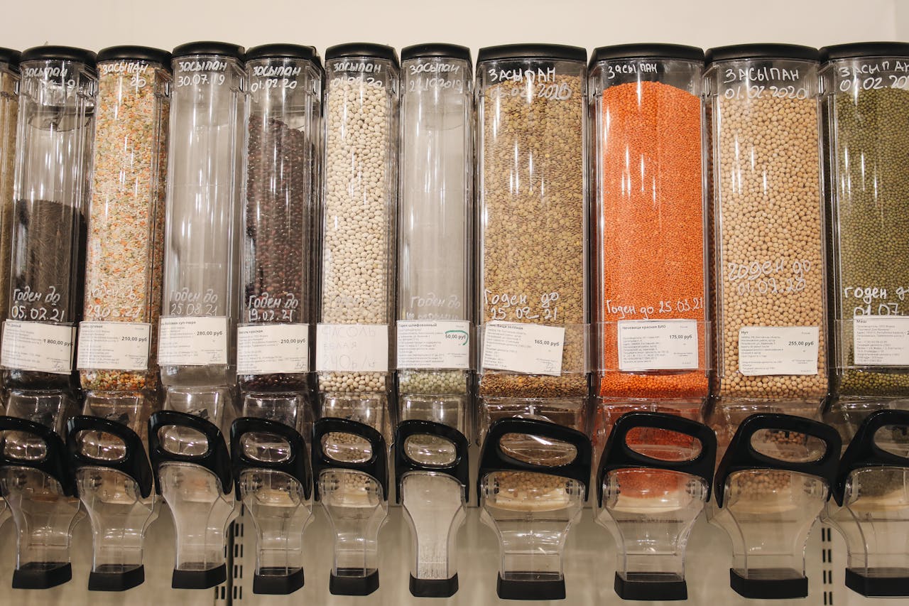 Bulk food dispensers filled with grains, legumes, and lentils, clear containers with scoops, labels and prices visible