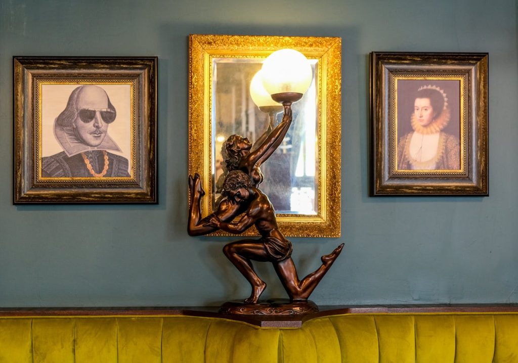 Vintage-style interior with framed portraits on a dark green wall, ornate gold frames surrounding artwork of historical figures, unique bronze statue lamp of a kneeling figure