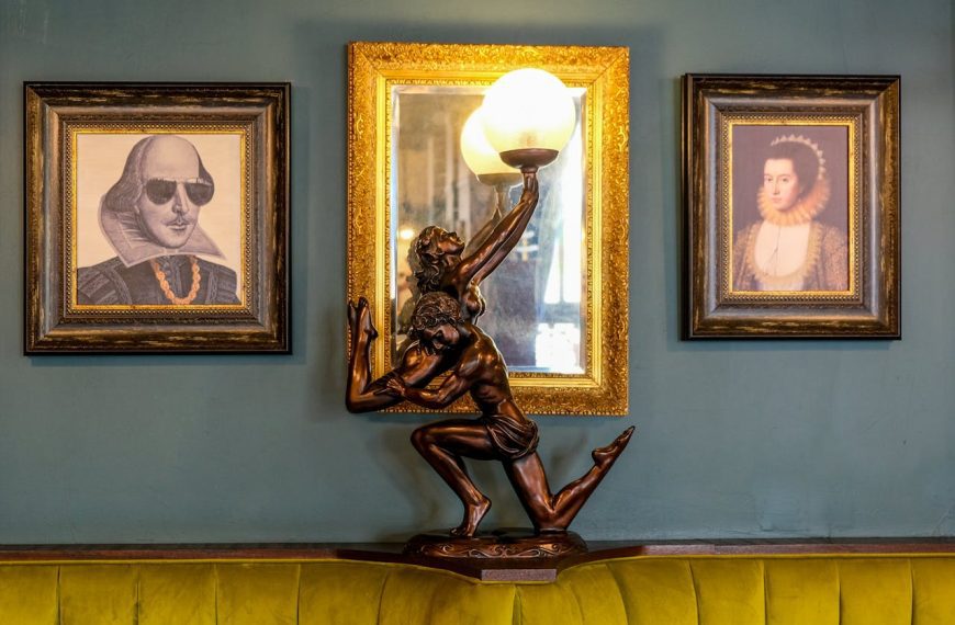 Vintage-style interior with framed portraits on a dark green wall, ornate gold frames surrounding artwork of historical figures, unique bronze statue lamp of a kneeling figure