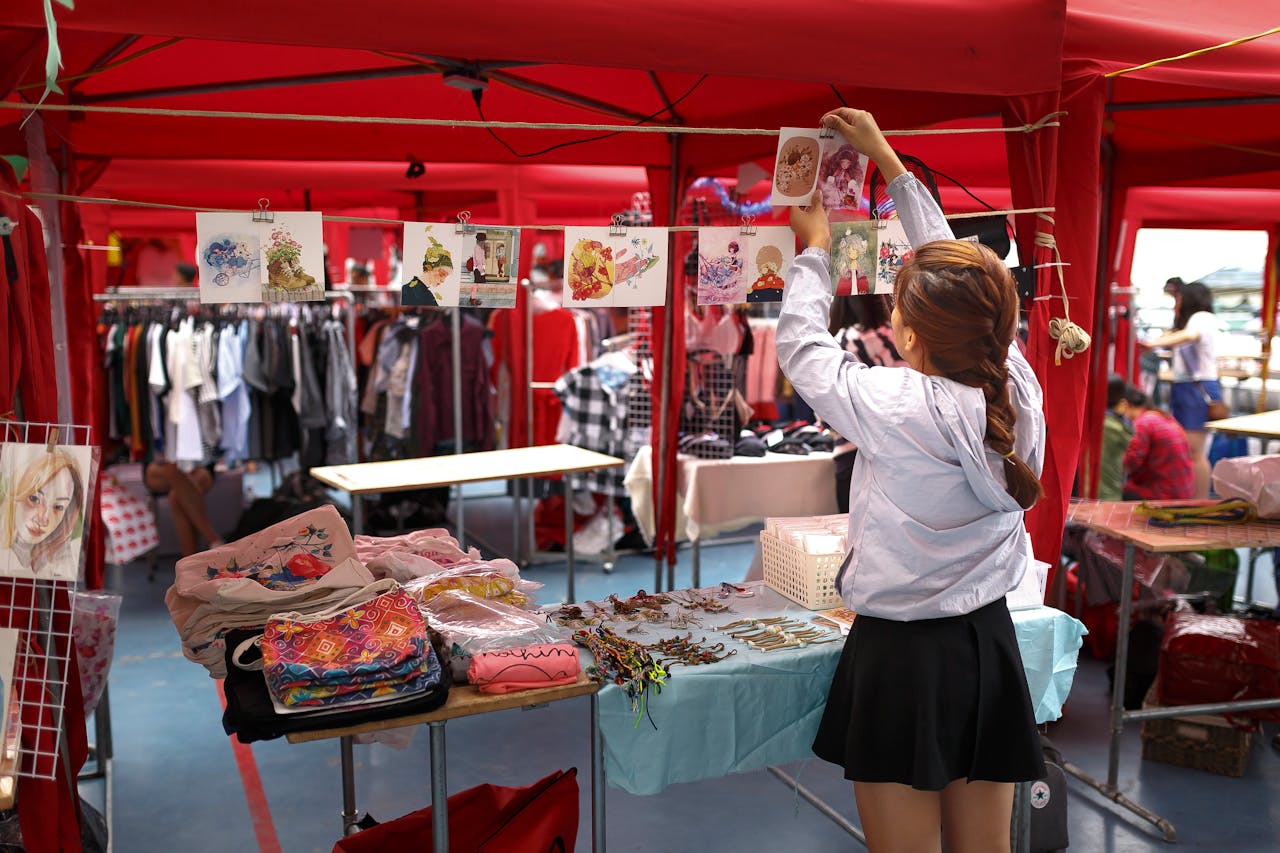 Woman hanging prints at a red market stall, table with handmade items, clothes and accessories in background