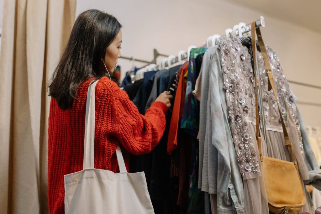 Woman in red sweater browsing dresses on a clothing rack, tote bag on shoulder, indoor store setting