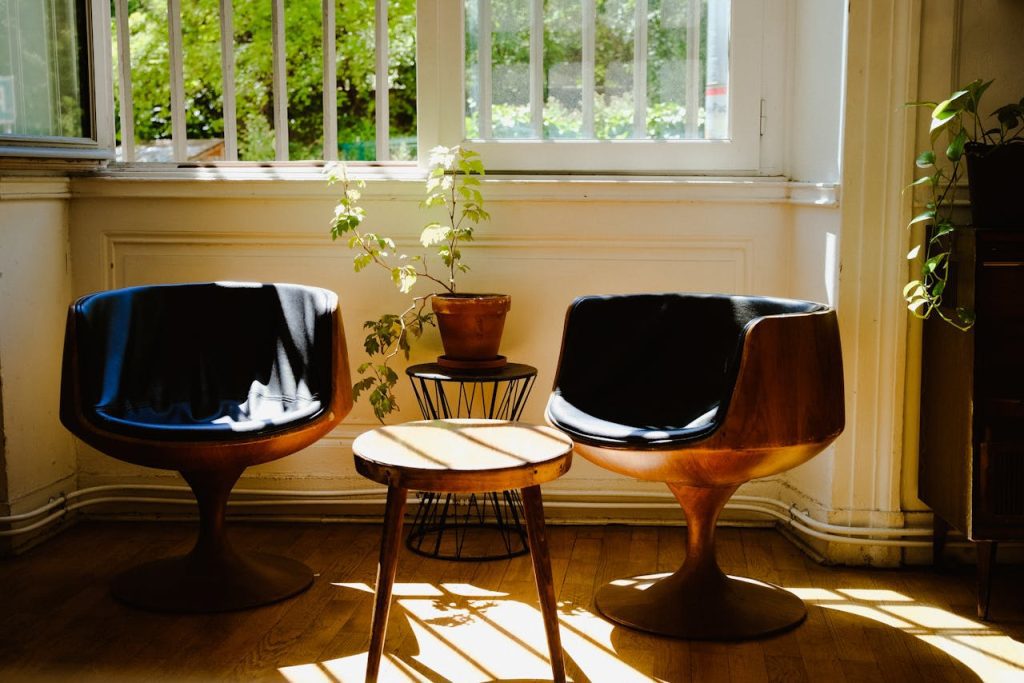 Two modern wooden chairs with black cushions, small round table in between, potted plant, sunlit room with open window