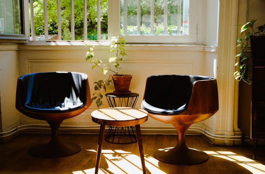 Two modern wooden chairs with black cushions, small round table in between, potted plant, sunlit room with open window
