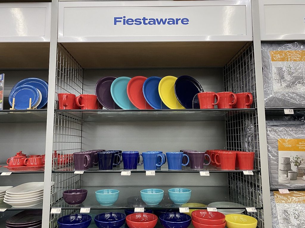 Colorful display of plates, bowls, and mugs, neatly arranged on shelves, labeled “FINGERBOWLS,” inside a store or kitchenware section