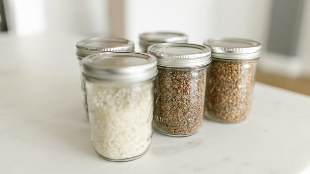 Glass jars with metal lids, filled with grains and seeds, arranged on white countertop, soft natural lighting