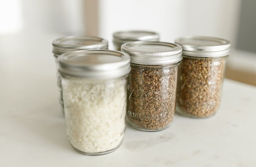 Glass jars with metal lids, filled with grains and seeds, arranged on white countertop, soft natural lighting