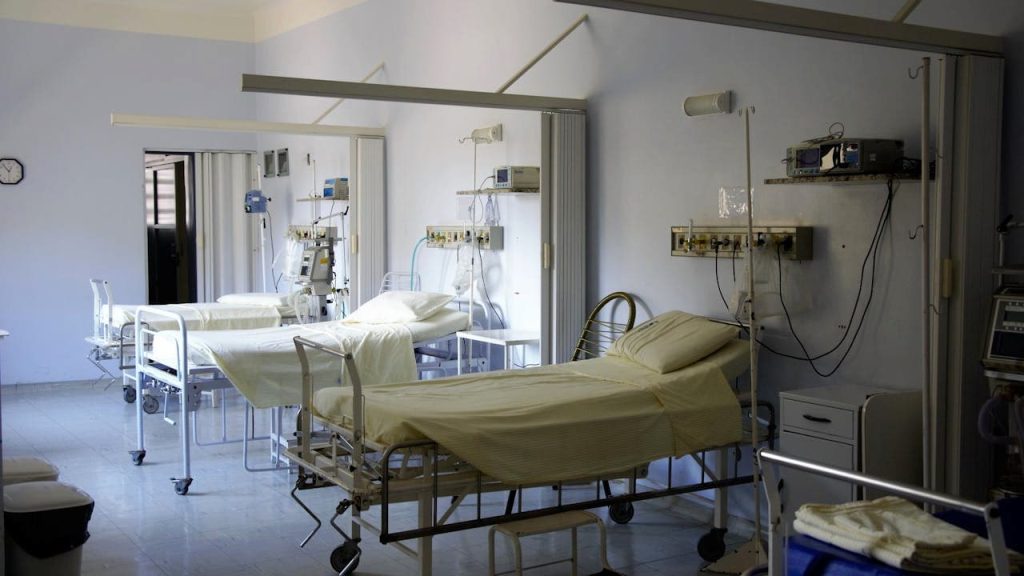 A quiet hospital ward with multiple empty beds, medical equipment, white walls, and dim lighting, creating a sterile and clinical atmosphere