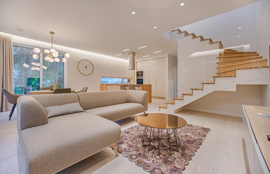 Modern open-concept living room with a beige sectional sofa, round coffee table on a patterned rug, floating wooden staircase with white accents, warm ambient lighting