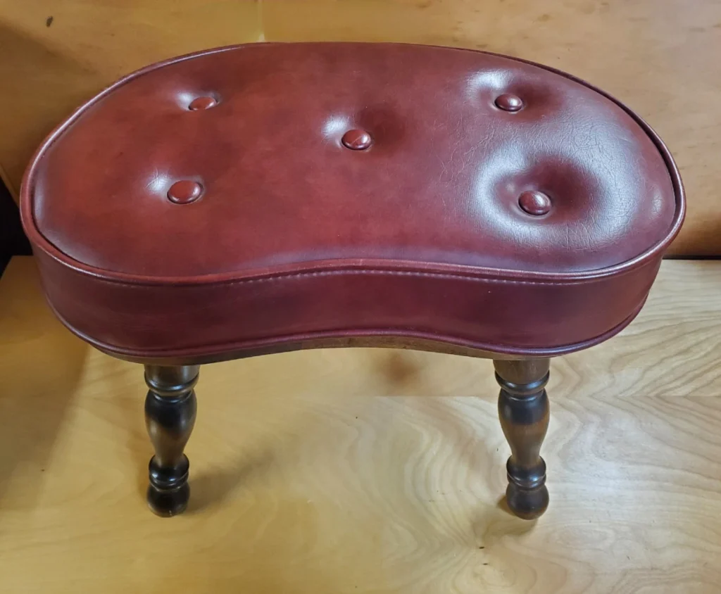 Vintage kidney-shaped stool, red leather upholstery with button tufting, dark wooden legs with a curved design, placed on a wooden surface, antique or classic furniture piece