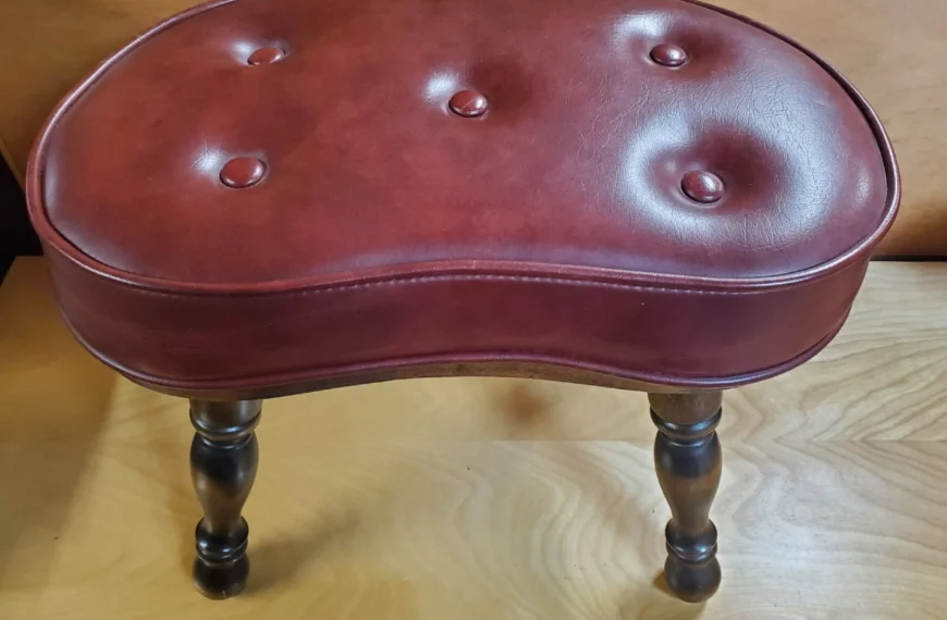 Vintage kidney-shaped stool, red leather upholstery with button tufting, dark wooden legs with a curved design, placed on a wooden surface, antique or classic furniture piece