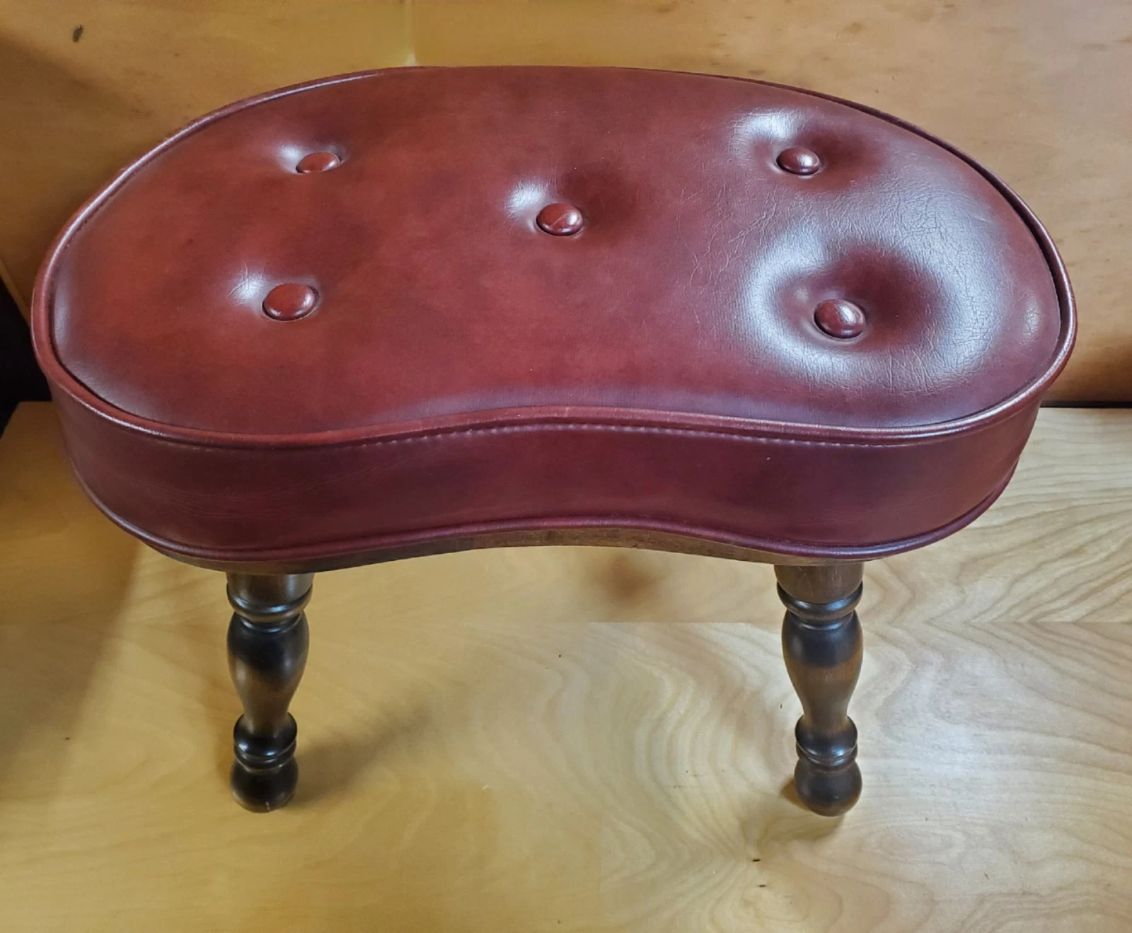 Vintage kidney-shaped stool, red leather upholstery with button tufting, dark wooden legs with a curved design, placed on a wooden surface, antique or classic furniture piece