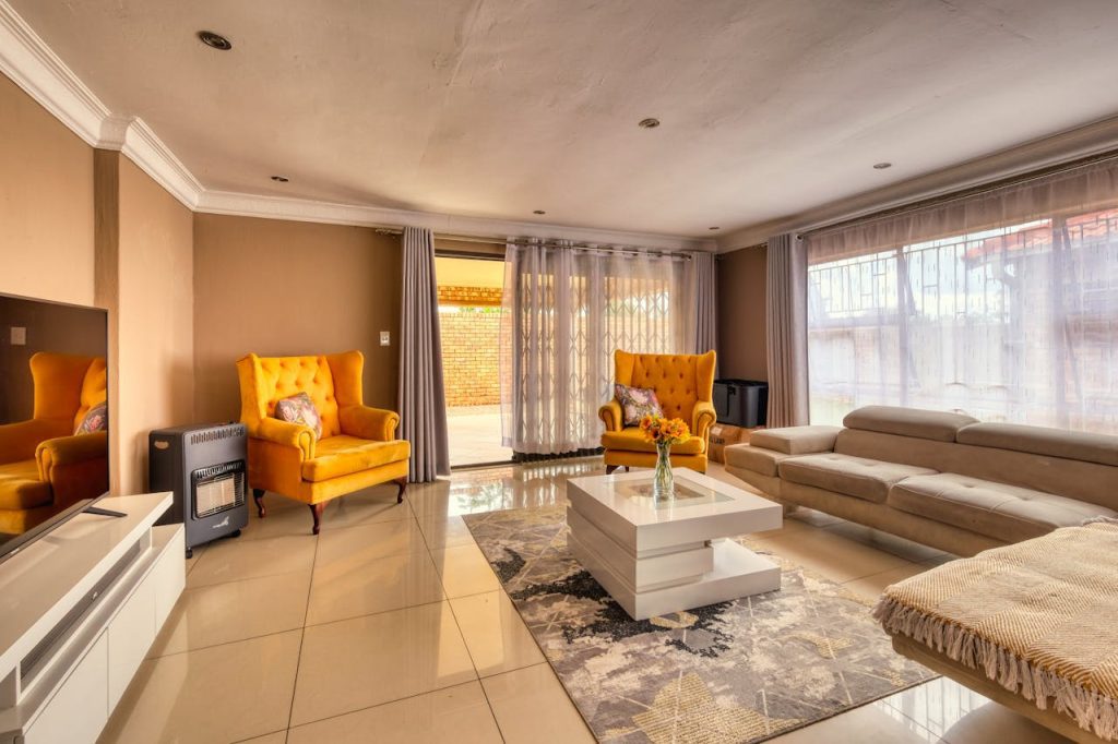 Modern living room, warm lighting, two mustard-yellow armchairs, large windows with sheer curtains, beige sectional sofa, glass-top coffee table with flowers