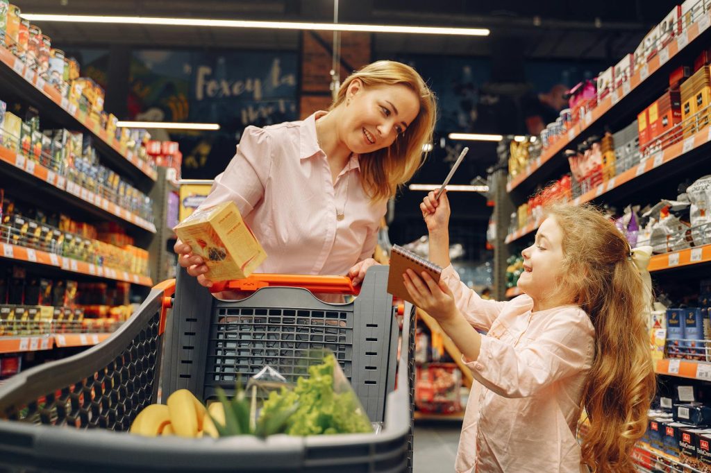Woman and young girl grocery shopping, standing beside a cart, smiling, checking items on a shopping list