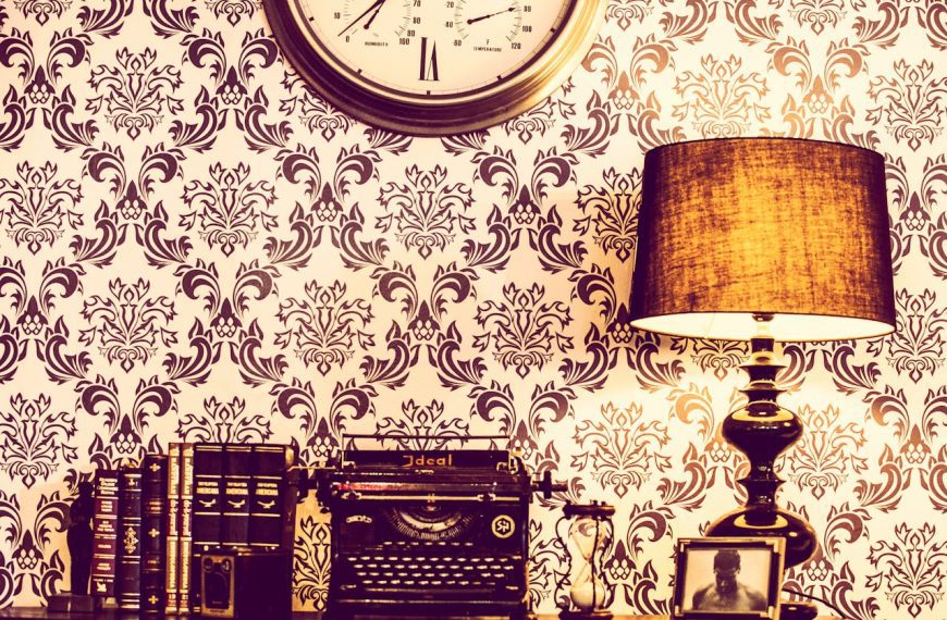 Vintage desk setup with typewriter, table lamp, books, ornate wallpaper background, round wall clock above