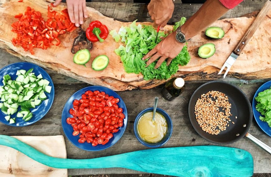Hands preparing a fresh salad, wooden table with chopped vegetables, blue plates filled with tomatoes, cucumbers, and dressing, outdoor cooking setting, natural and rustic food preparation