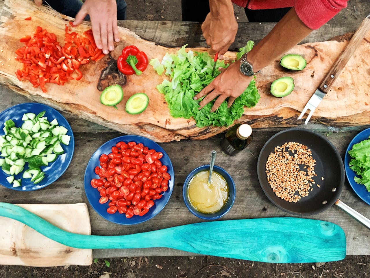 Hands preparing a fresh salad, wooden table with chopped vegetables, blue plates filled with tomatoes, cucumbers, and dressing, outdoor cooking setting, natural and rustic food preparation