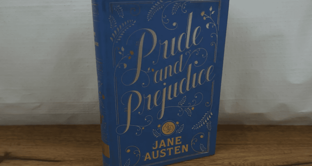 Blue hardcover of “Pride and Prejudice” by Jane Austen, gold embossed lettering and floral design, upright on wooden surface