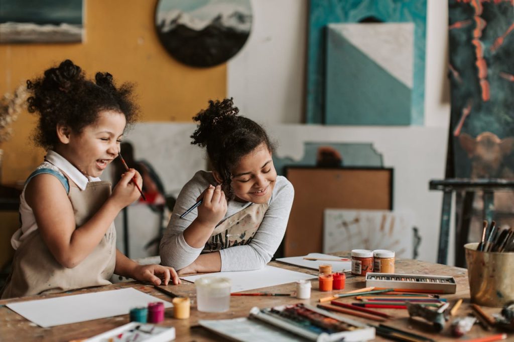 Two young girls with curly hair, wearing aprons, sitting at a table, painting and smiling, surrounded by art supplies, in a creative studio with colorful artwork on the walls