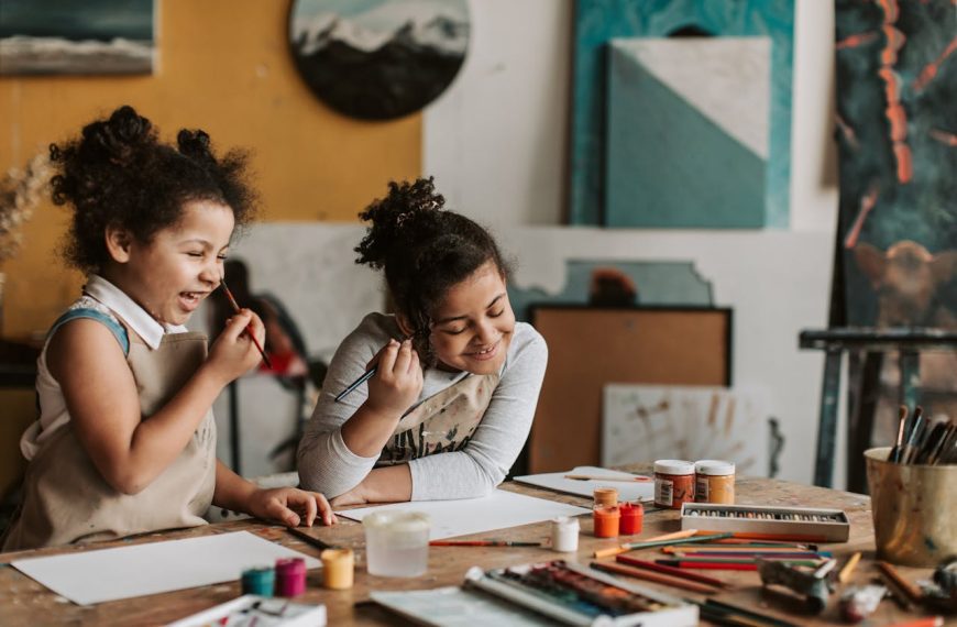 Two young girls with curly hair, wearing aprons, sitting at a table, painting and smiling, surrounded by art supplies, in a creative studio with colorful artwork on the walls