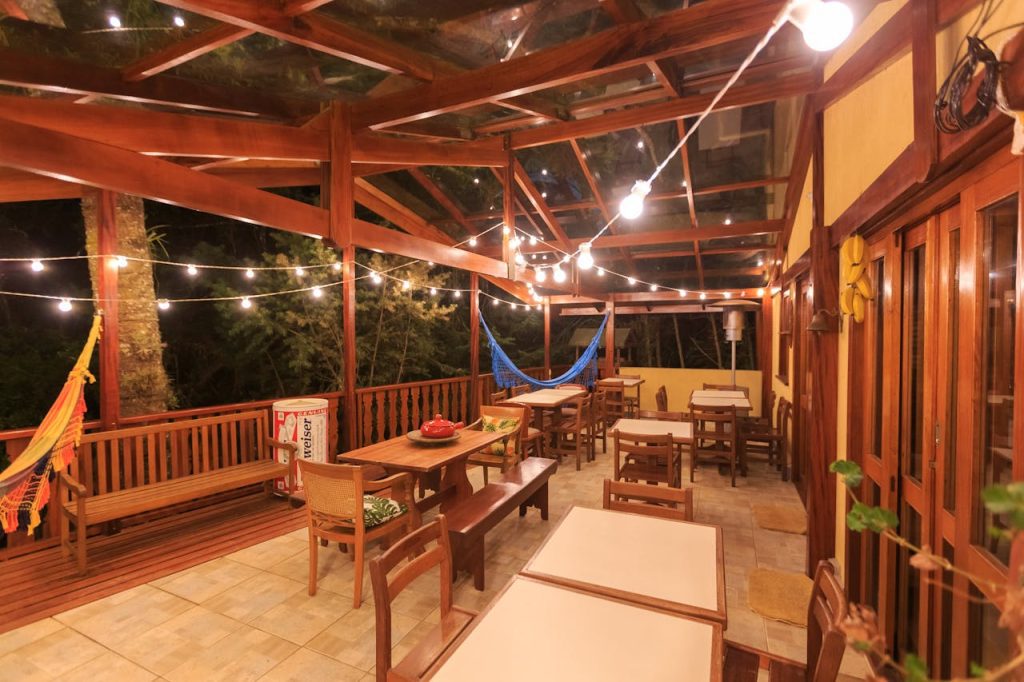 Wooden outdoor patio with string lights, dining tables, bench seating, hammock in back, warm night setting, tiled floor